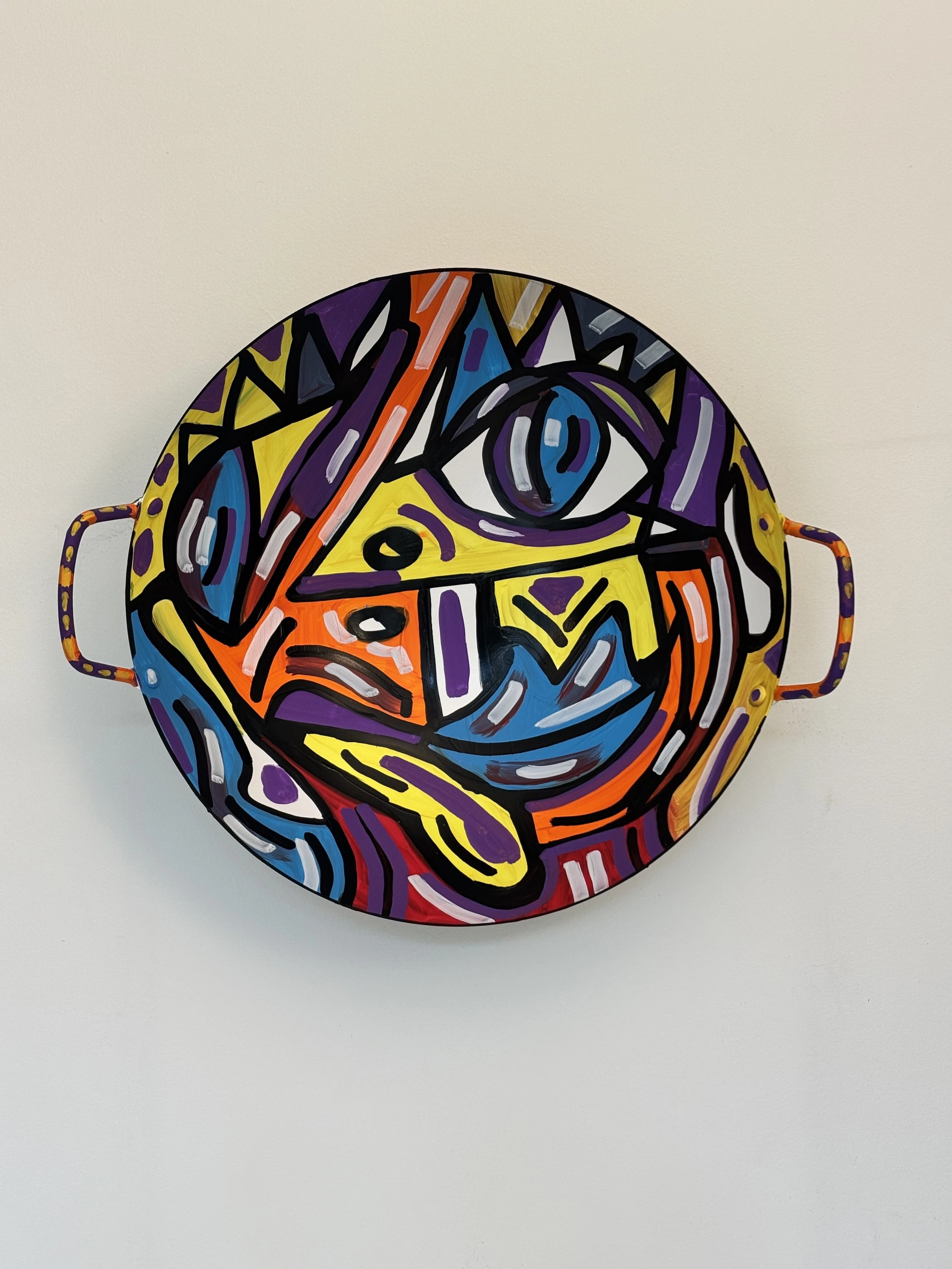 Cuenco con asas colgado en la pared, con un rostro de un solo ojo azul y una lengua amarilla, en violeta, naranja, azul y blanco entre líneas negras. Bowl. Acrílico sobre hierro, 38 cm de diámetro, 2025. Caracas. Nicolás Lapadula