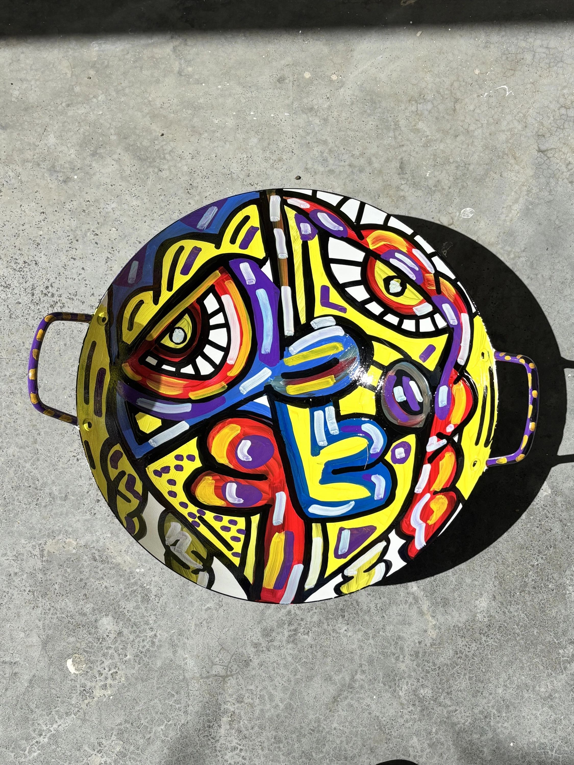 Cuenco de hierro con asas laterales, visto desde arriba, con un rostro dividido en amarillo, azul, rojo, naranja y violeta. Bowl. Acrílico sobre hierro, 38 cm de diámetro, 2025. Caracas. Nicolás Lapadula