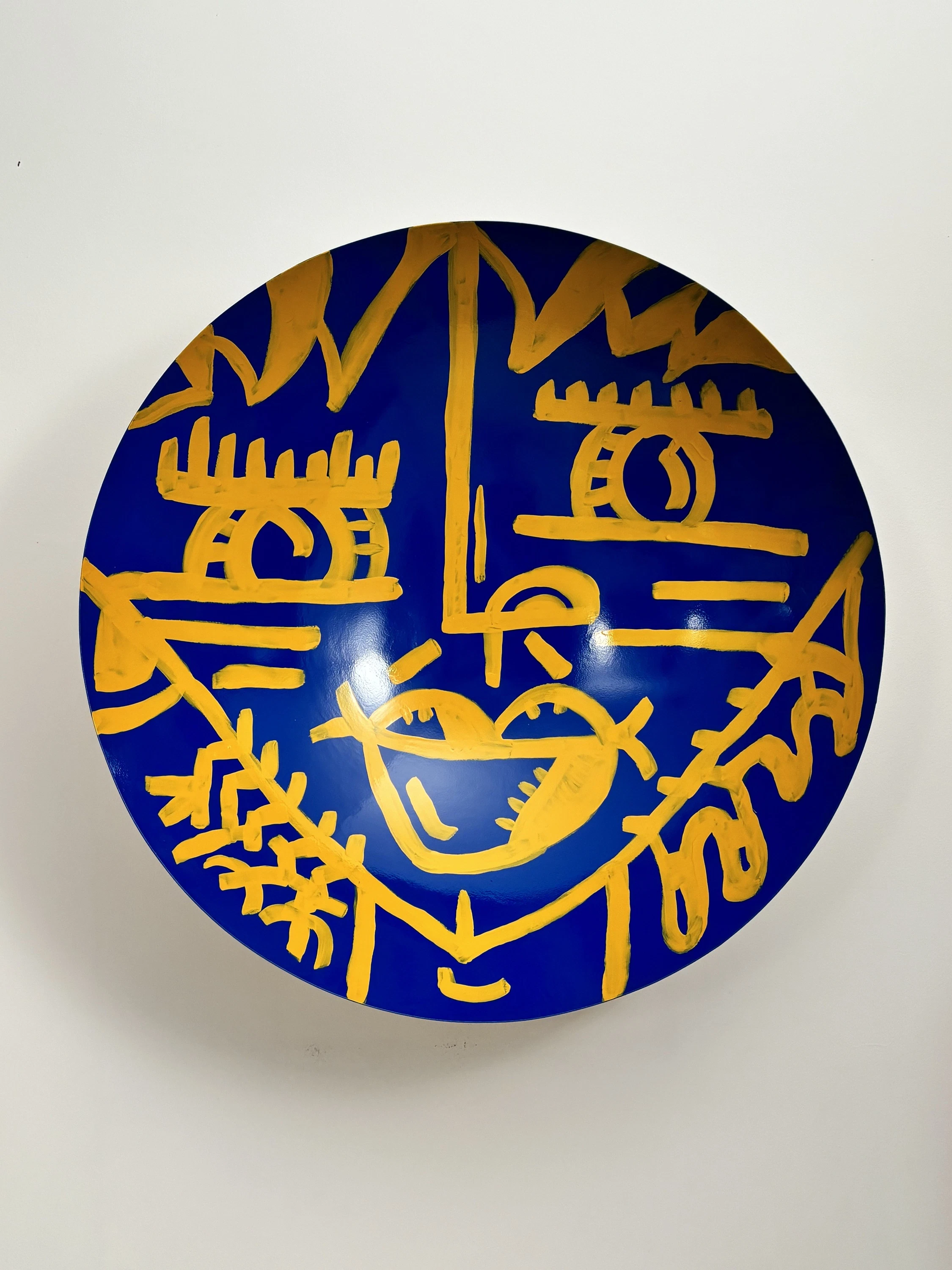 Cuenco hondo con un rostro de ojos de pestañas largas y boca en forma de corazón, en líneas amarillas sobre azul intenso. Bowl. Acrílico sobre hierro, 73 cm de diámetro x 24 cm de profundidad, 2025. Caracas. Nicolás Lapadula