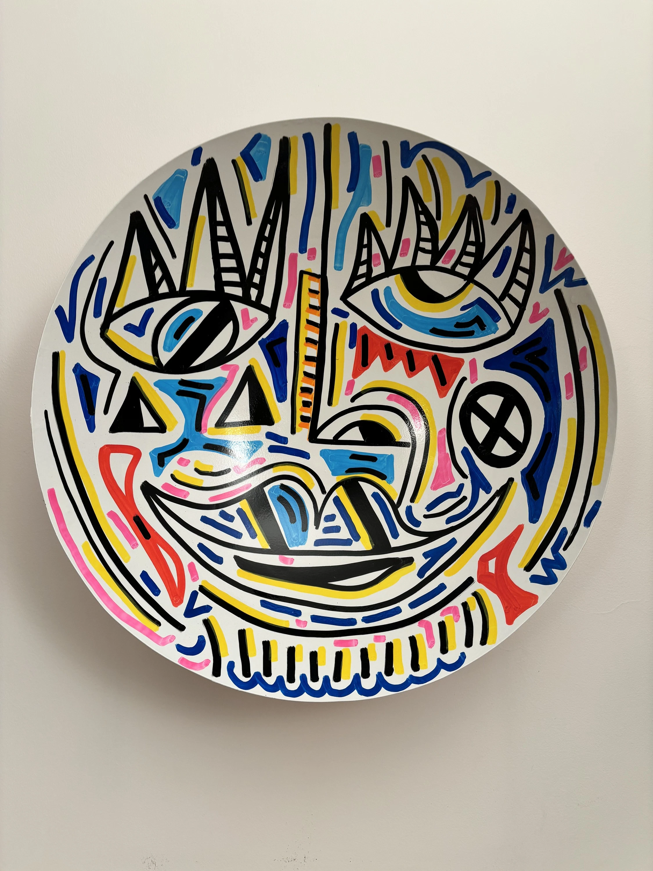 Cuenco blanco colgado en la pared, con un rostro sonriente de ojos con pestañas puntiagudas, en líneas negras, amarillas, azules y rosadas. Bowl. Acrílico sobre hierro, 50 cm de diámetro, 2025. Caracas. Nicolás Lapadula