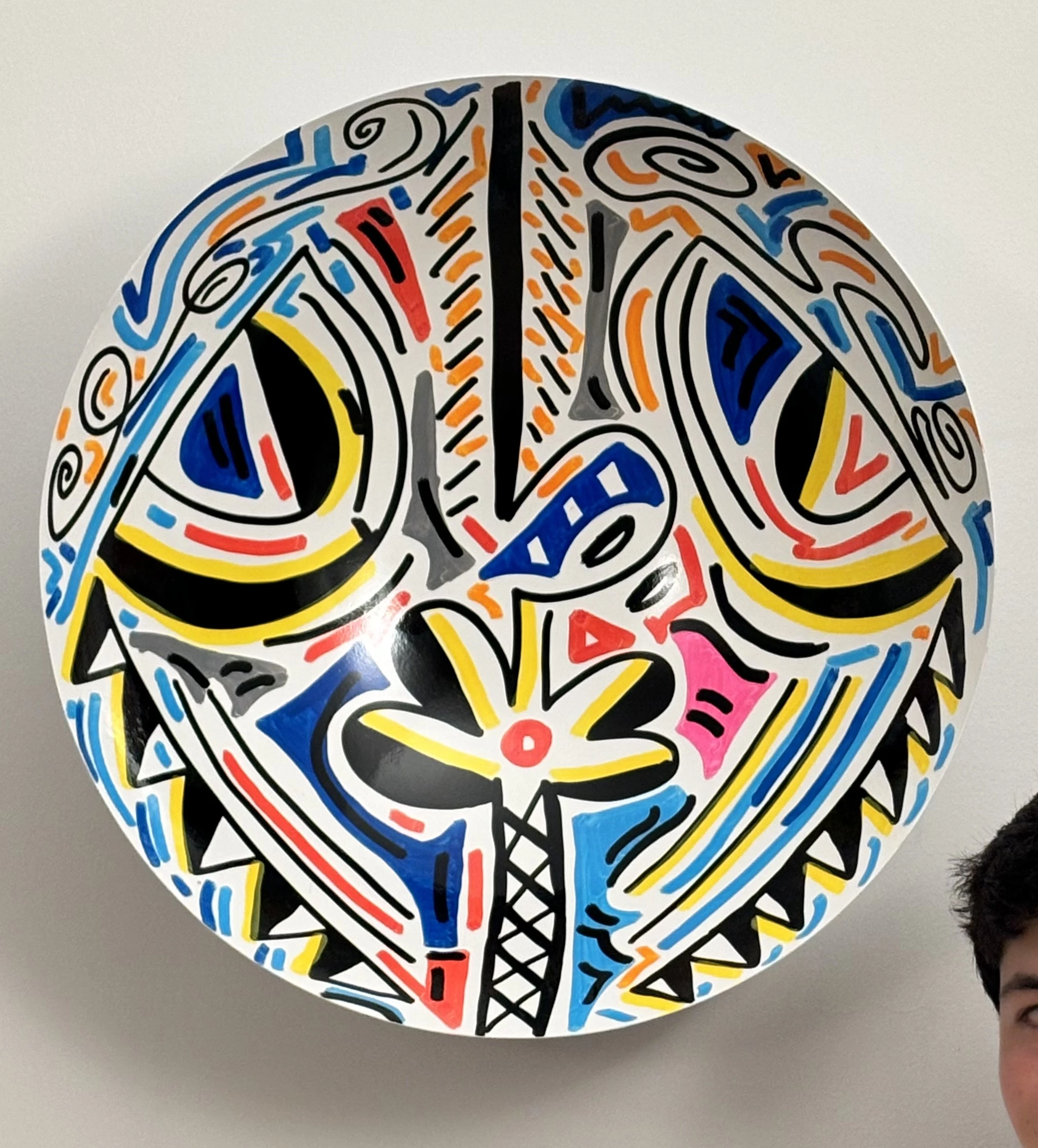 Cuenco blanco colgado en la pared, con una máscara de dos ojos grandes de trazo negro y amarillo, dientes dentados y detalles azules, naranjas y rosados. Bowl. Acrílico sobre hierro, 50 cm de diámetro, 2025. Caracas. Nicolás Lapadula