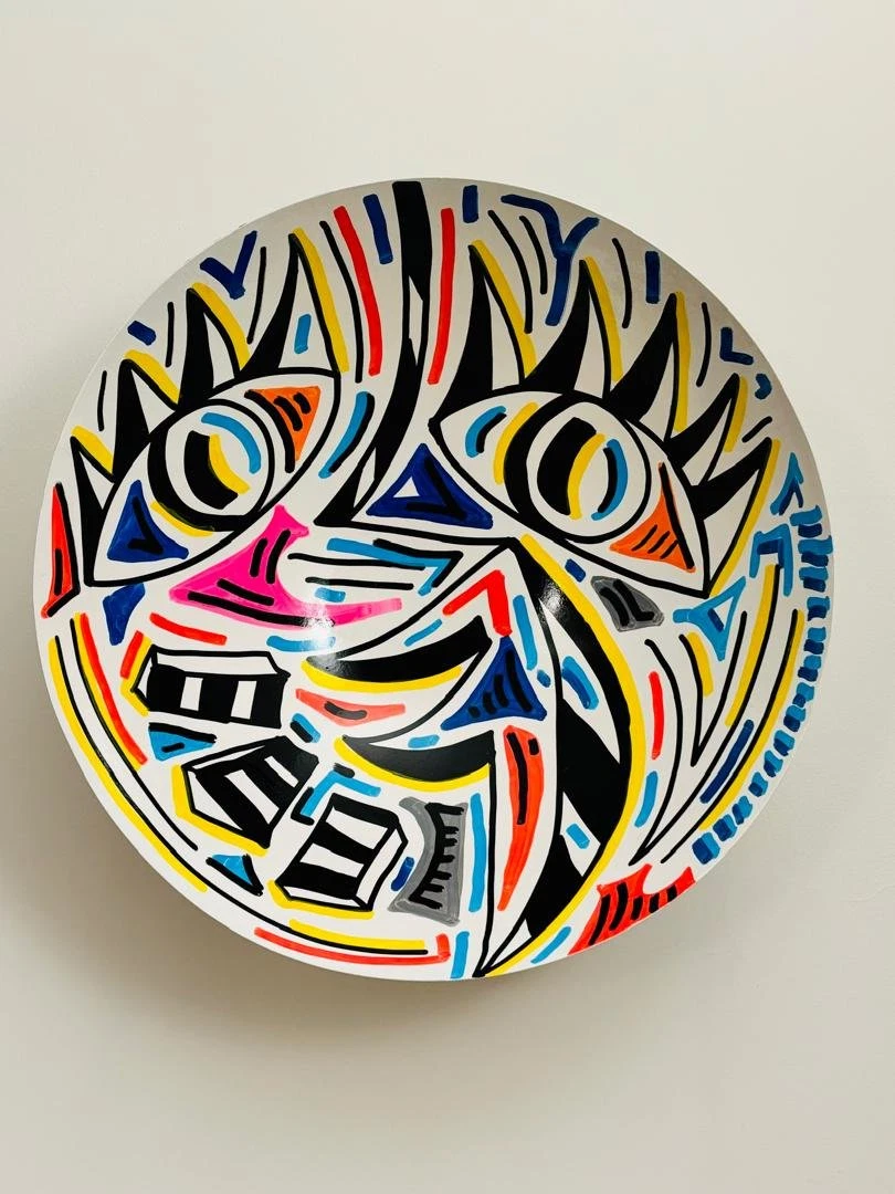 Cuenco blanco de pared con dos ojos de pestañas negras, una nariz rosada y una boca de bloques negros y blancos, entre trazos azules, rojos y amarillos. Bowl. Acrílico sobre hierro, 50 cm de diámetro, 2025. Caracas. Nicolás Lapadula