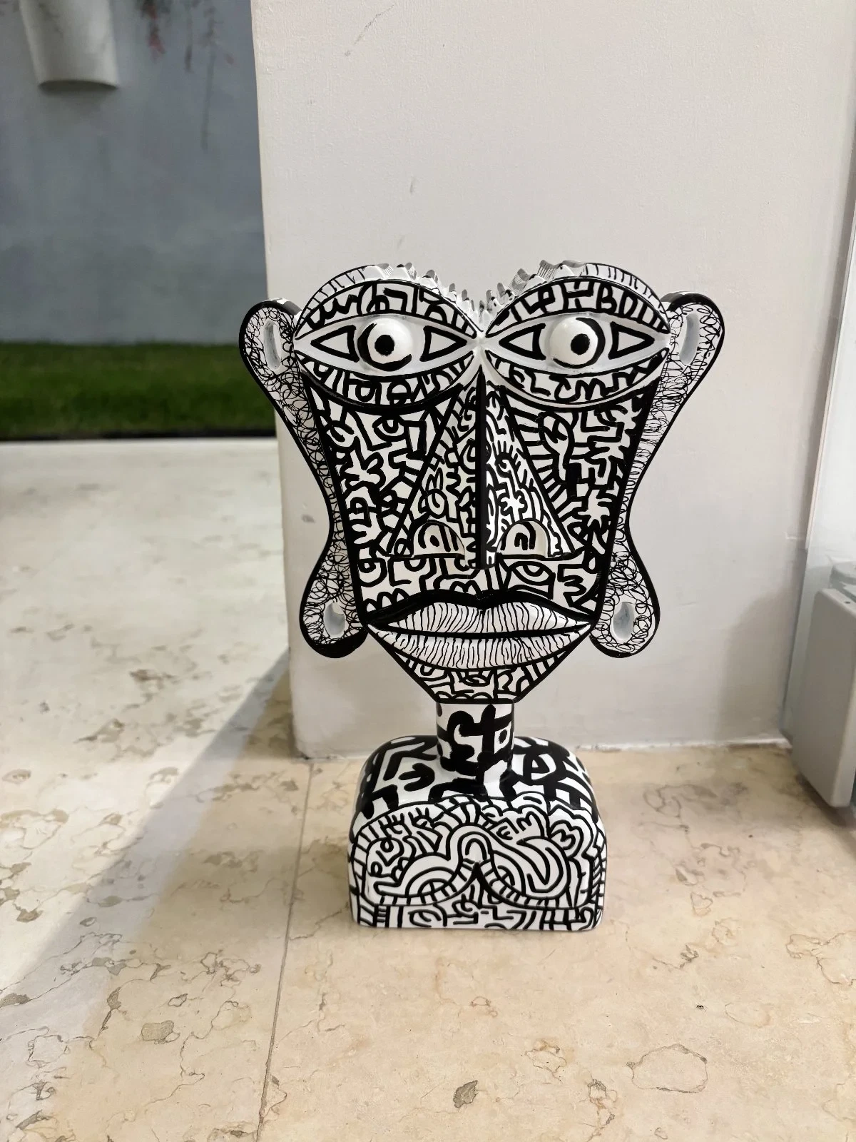 Busto blanco de rostro plano con ojos grandes y dibujos lineales negros, apoyado en un suelo de mármol junto a una columna. Busto. Acrílico sobre resina, 28 x 18 x 10 cm, 2026. Caracas. Nicolás Lapadula
