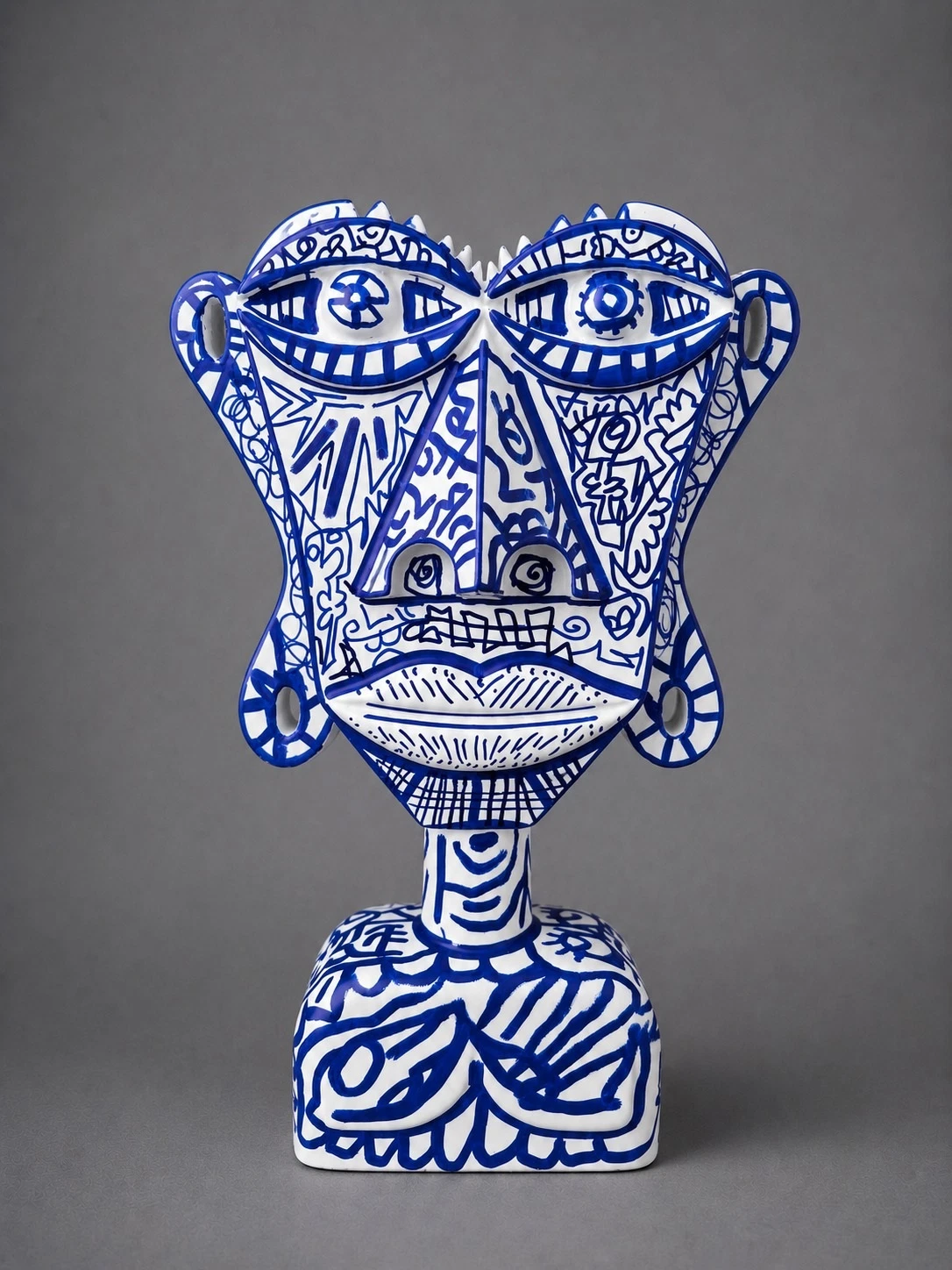Busto blanco de rostro plano y ojos grandes, cubierto de dibujos lineales en azul cobalto, sobre fondo gris. Busto. Acrílico sobre resina, 28 x 18 x 10 cm, 2026. Caracas. Nicolás Lapadula