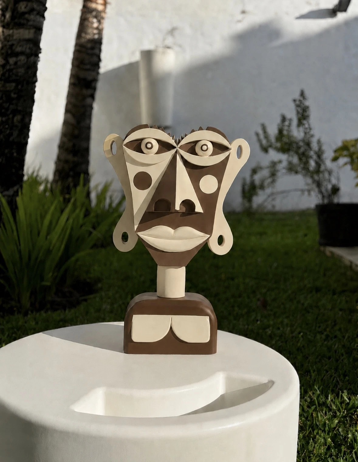 Busto de rostro frontal con ojos redondos y orejas en forma de asa, en marrón y crema, sobre un pedestal blanco en un jardín. Busto. Acrílico sobre resina, 28 x 18 x 10 cm, 2026. Caracas. Nicolás Lapadula