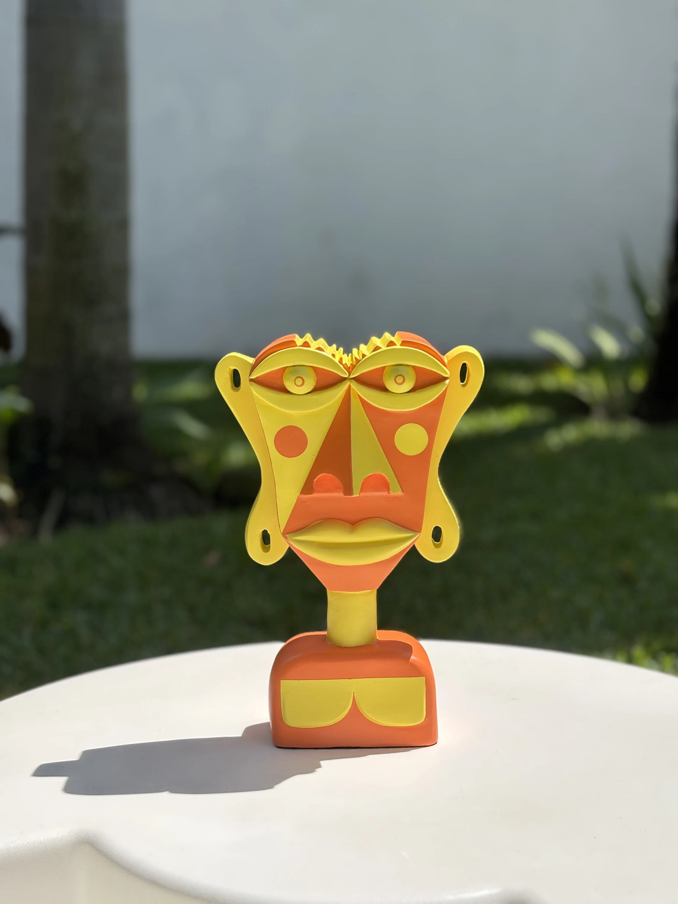 Busto de rostro plano pintado en amarillo y naranja, con círculos en las mejillas, sobre una mesa blanca al aire libre. Busto. Acrílico sobre resina, 28 x 18 x 10 cm, 2026. Caracas. Nicolás Lapadula