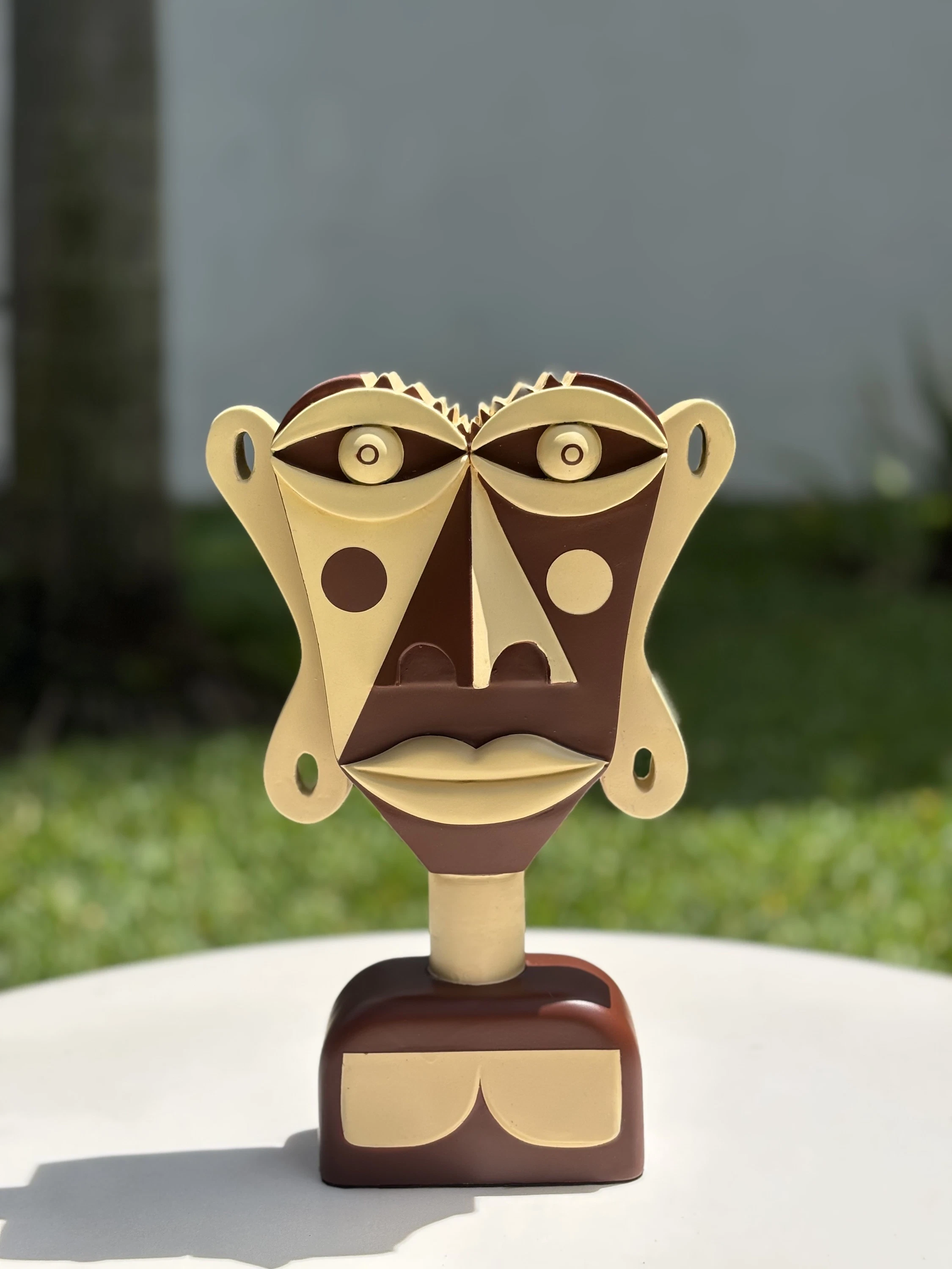 Busto de rostro plano pintado en marrón oscuro y crema, con círculos en las mejillas, sobre una mesa blanca al aire libre. Busto. Acrílico sobre resina, 28 x 18 x 10 cm, 2026. Caracas. Nicolás Lapadula