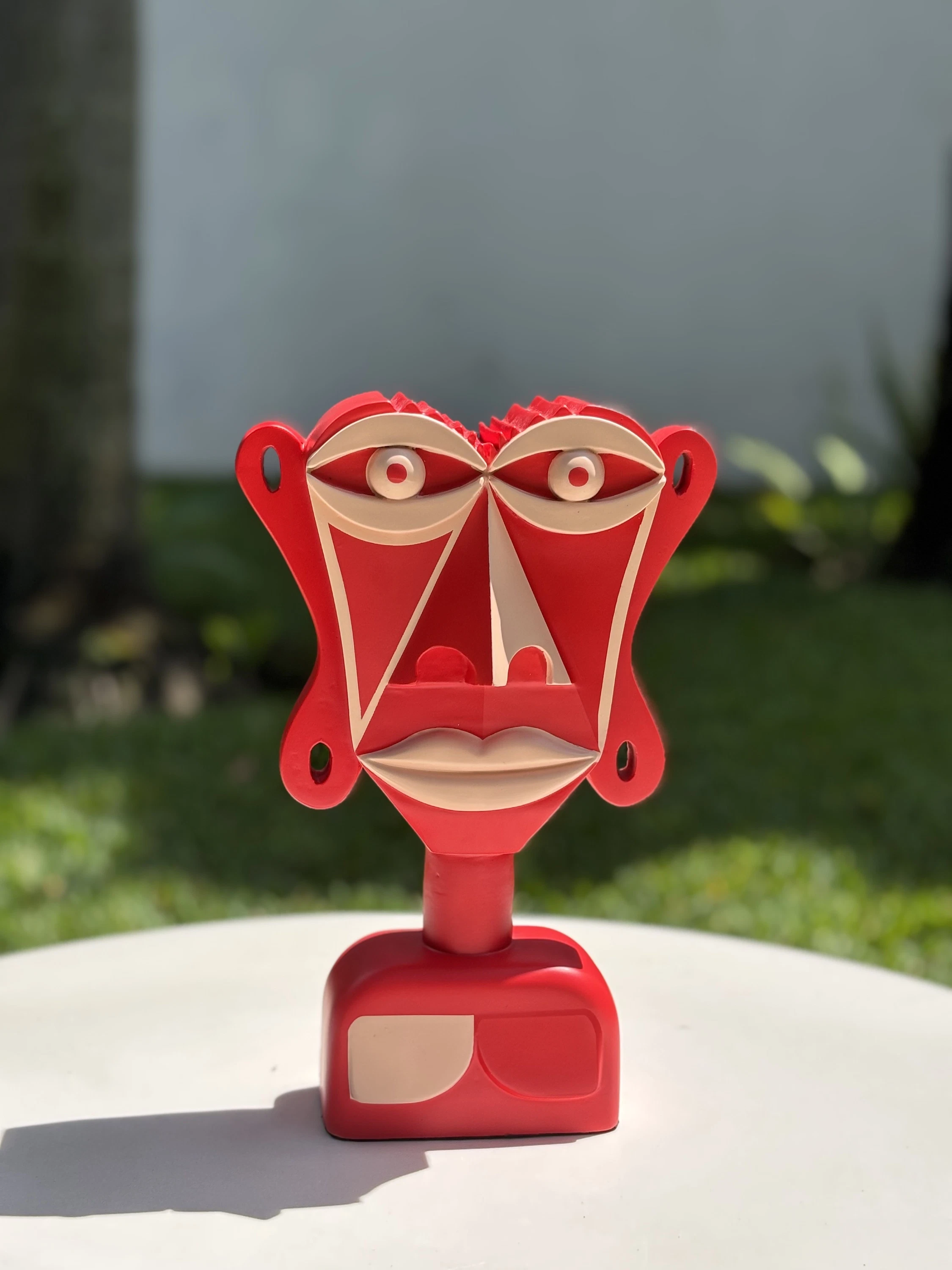 Busto de rostro plano pintado en rojo con líneas color crema alrededor de los ojos y la nariz, sobre una mesa blanca al aire libre. Busto. Acrílico sobre resina, 28 x 18 x 10 cm, 2026. Caracas. Nicolás Lapadula