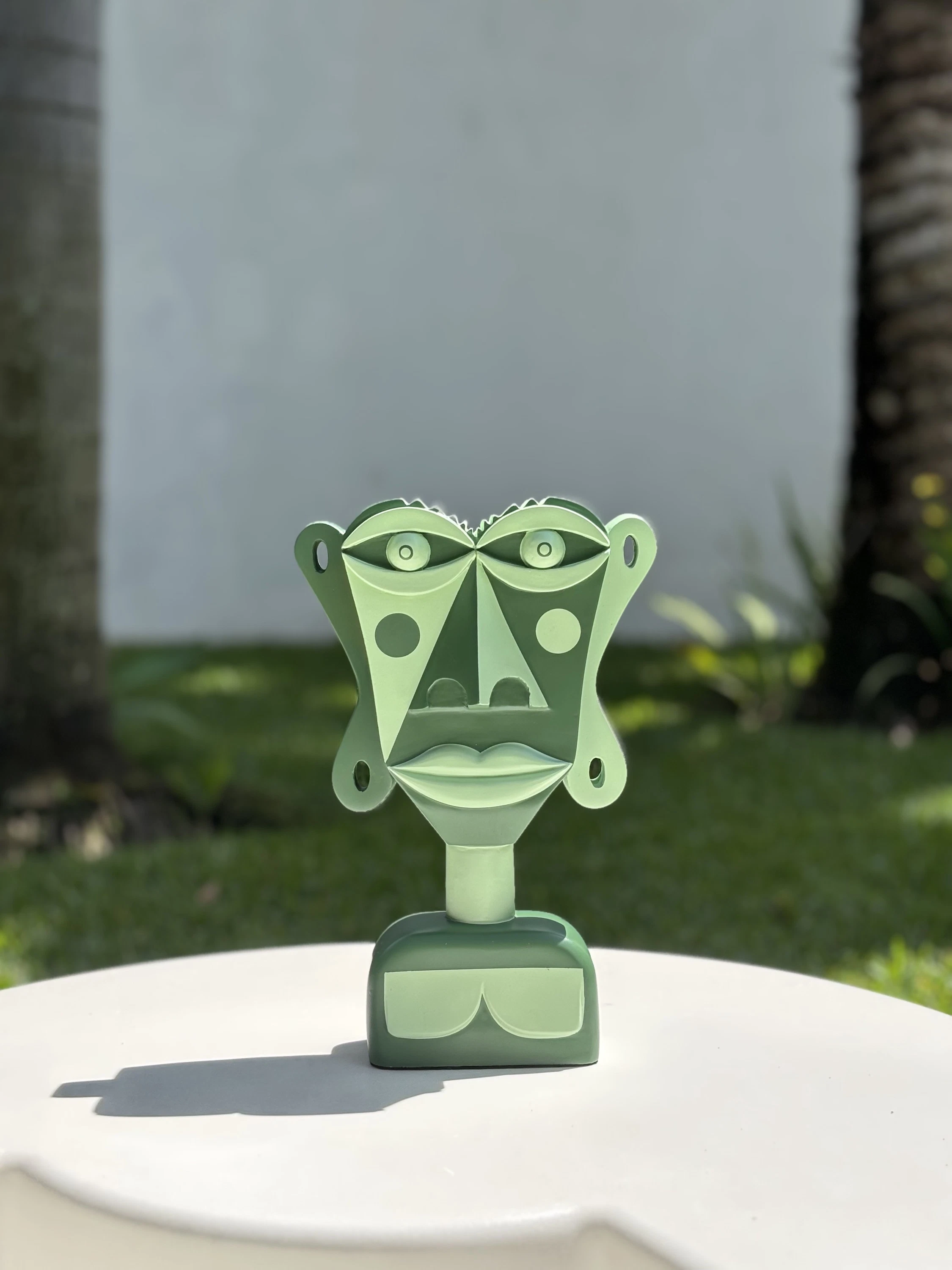 Busto de rostro plano pintado en tonos de verde, con círculos en las mejillas, sobre una mesa blanca al aire libre. Busto. Acrílico sobre resina, 28 x 18 x 10 cm, 2026. Caracas. Nicolás Lapadula