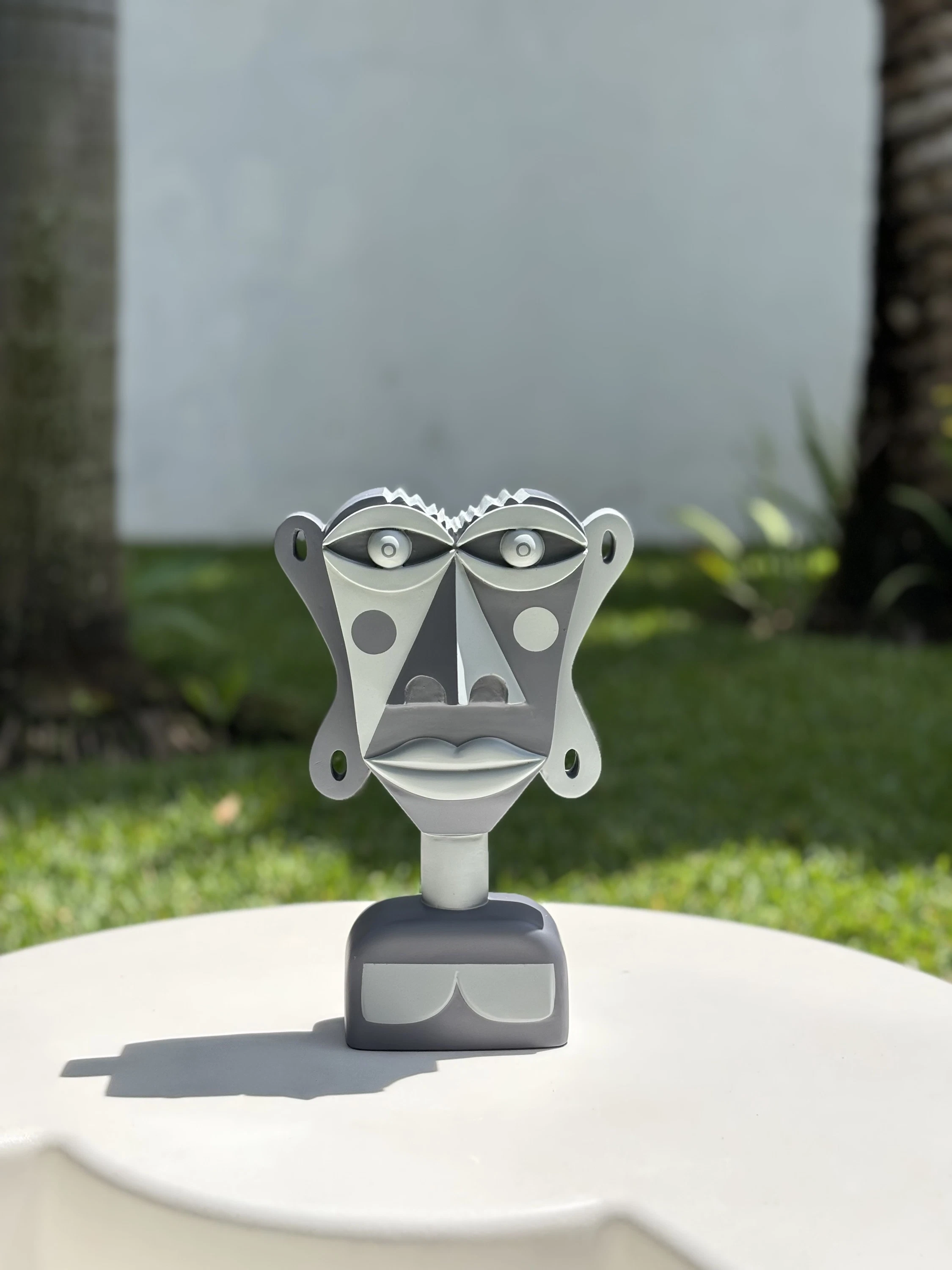 Busto de rostro plano pintado en tonos de gris, con círculos en las mejillas, sobre una mesa blanca al aire libre. Busto. Acrílico sobre resina, 28 x 18 x 10 cm, 2026. Caracas. Nicolás Lapadula