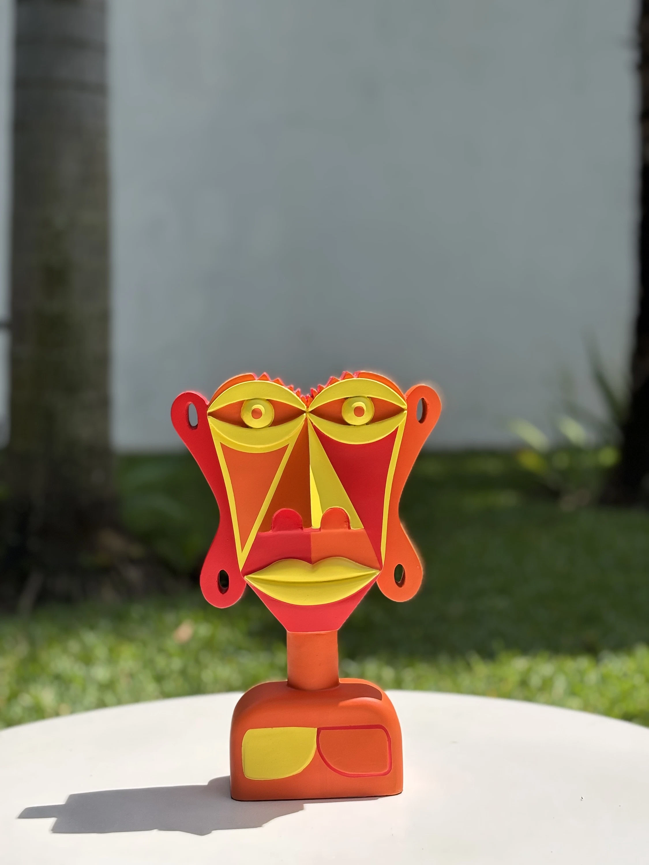 Busto de rostro plano pintado en rojo, naranja y amarillo, con los labios y los ojos amarillos, sobre una mesa blanca al aire libre. Busto. Acrílico sobre resina, 28 x 18 x 10 cm, 2026. Caracas. Nicolás Lapadula