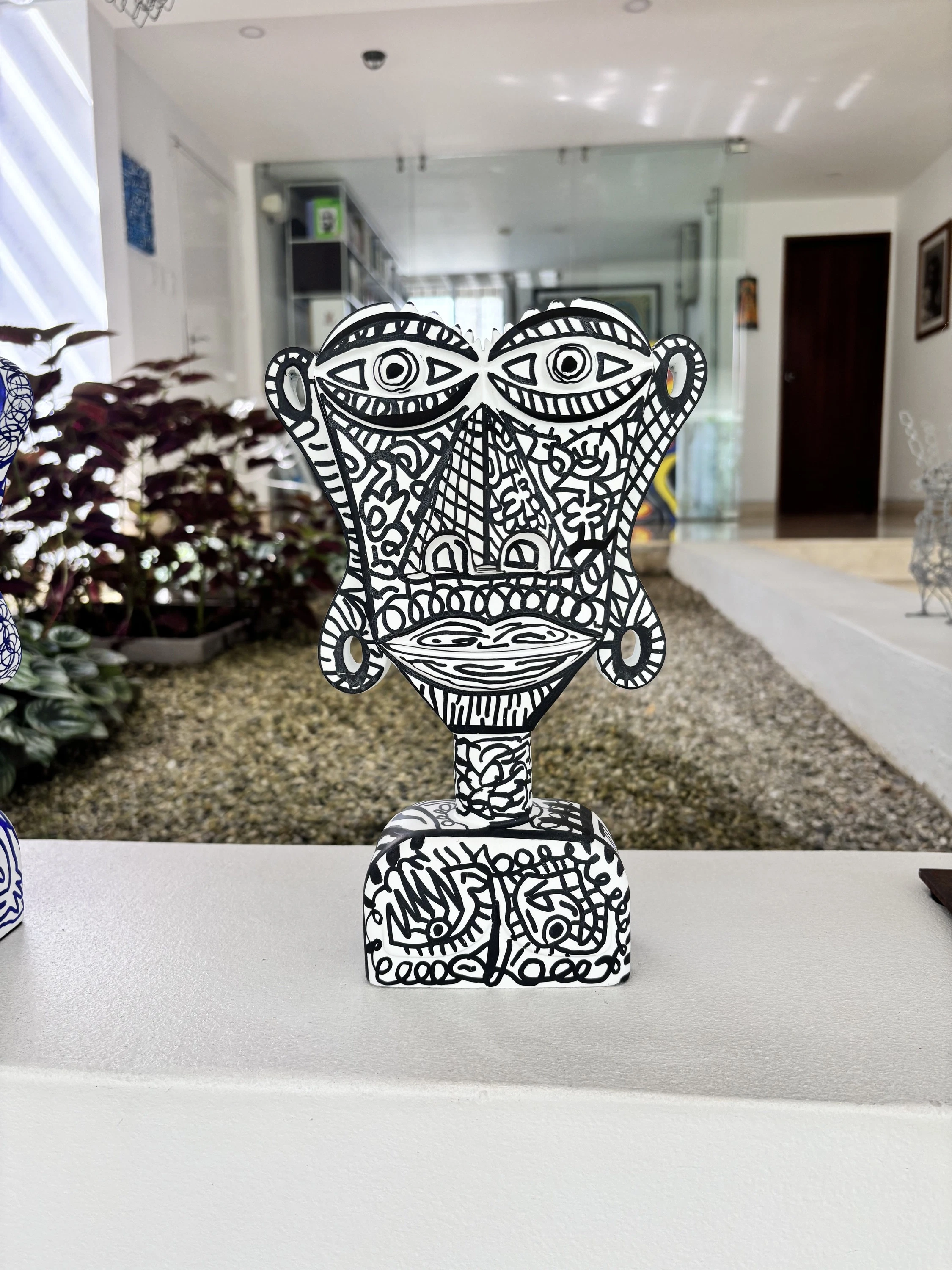 Busto blanco de rostro plano con ojos grandes y dibujos lineales negros muy finos, sobre una repisa en un pasillo con plantas. Busto. Acrílico sobre resina, 50 x 30 x 20 cm, 2026. Caracas. Nicolás Lapadula