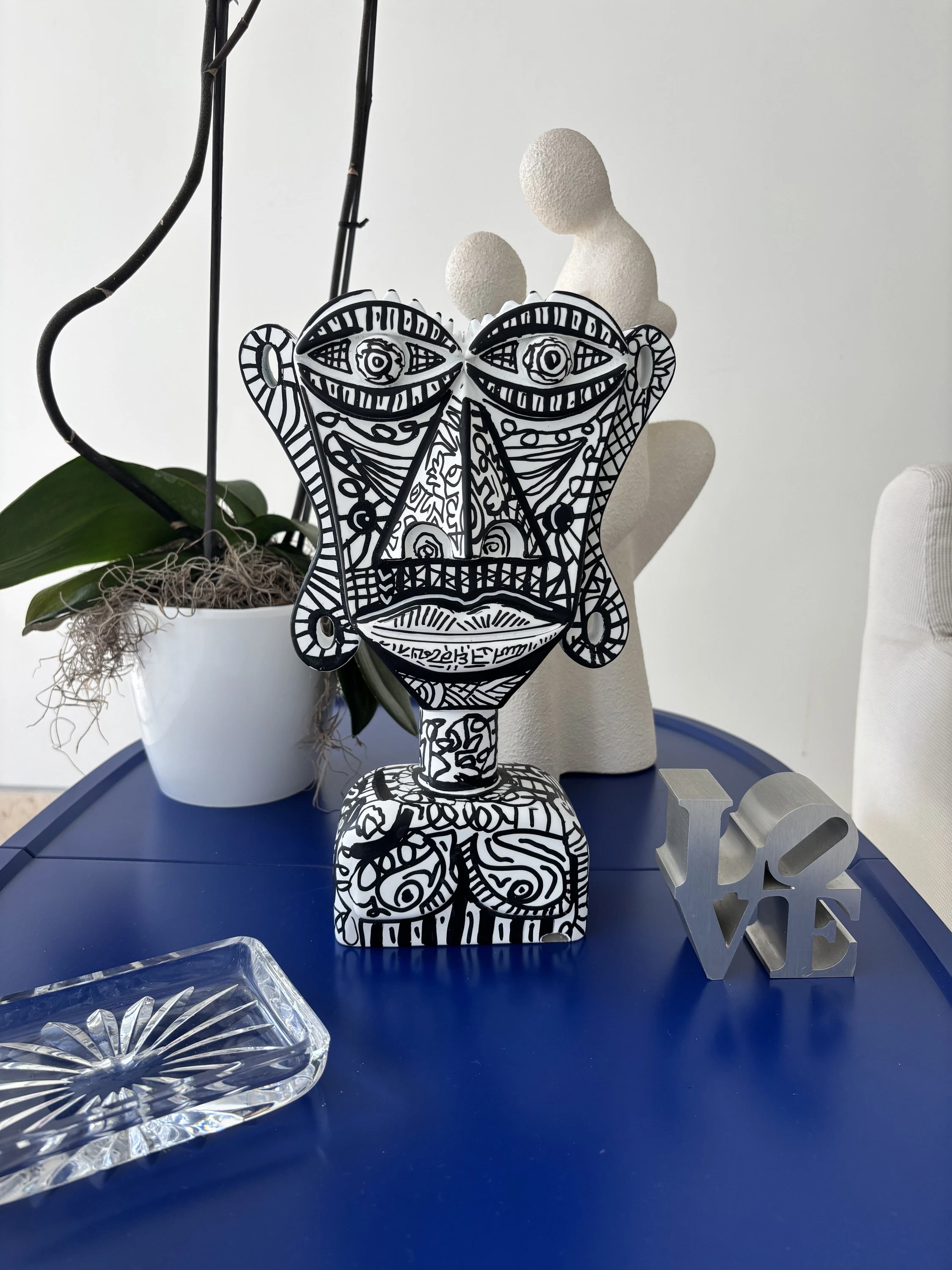 Busto blanco de rostro plano cubierto de dibujos lineales negros, sobre una mesa azul junto a una orquídea y una pequeña escultura plateada. Busto. Acrílico sobre resina, 28 x 18 x 10 cm, 2026. Caracas. Nicolás Lapadula