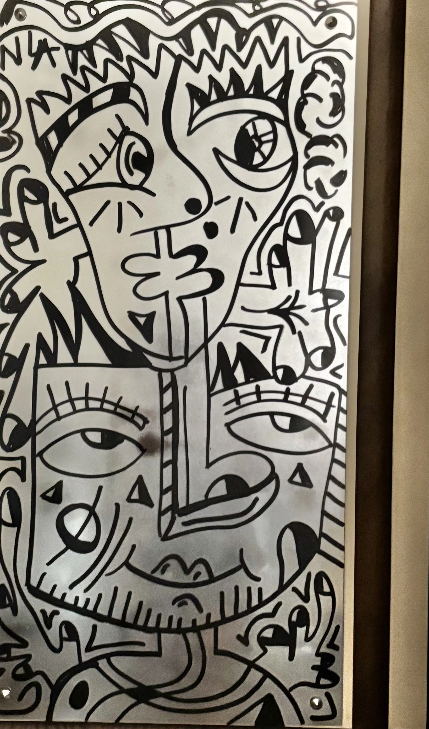 Tres rostros superpuestos de ojos grandes y cejas de rayas, dibujados en negro sobre una superficie plateada, con un marco oscuro a la derecha. Espejo. Acrílico sobre espejo, 89 x 55 cm, 2025. Madrid. Nicolás Lapadula