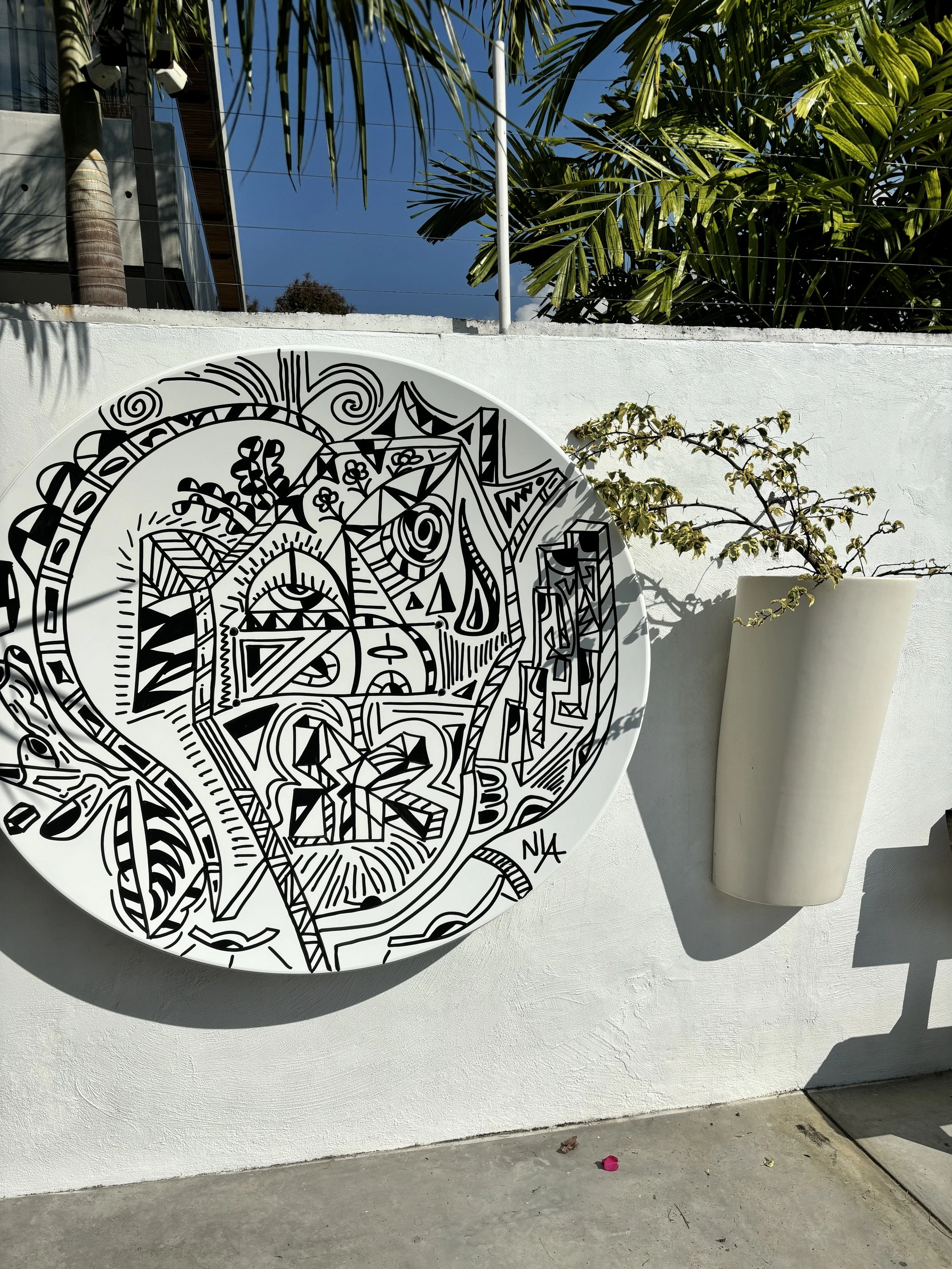 Disco blanco colgado de un muro blanco, con una composición de líneas, formas geométricas y rostros en negro, junto a una maceta con una planta. La Mujer. Acrílico sobre hierro, 150 x 165 cm, 2025. Caracas. Nicolás Lapadula