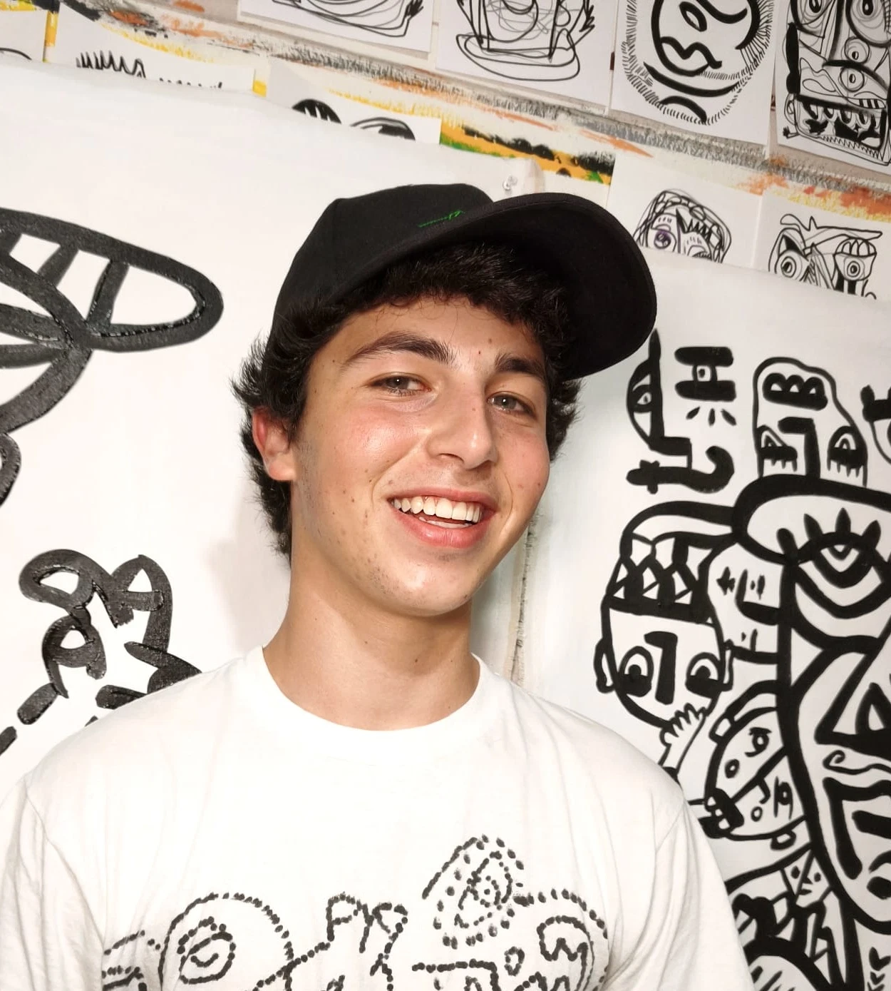 Nico sonriendo con gorra negra y una camiseta blanca cubierta de dibujos de línea, frente a sus pinturas murales en blanco y negro.