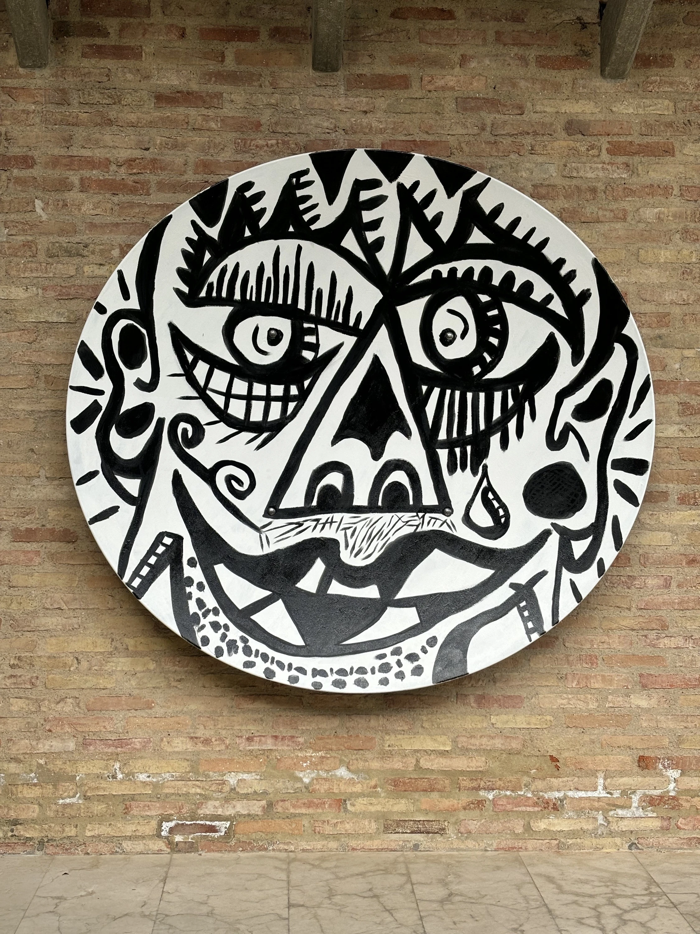 Disco blanco grande con un rostro negro de dientes, nariz triangular y ojos de pestañas, colgado de un muro de ladrillo. Rostro. Esmalte sobre plástico, 195 x 180 cm, 2024. Caracas. Nicolás Lapadula