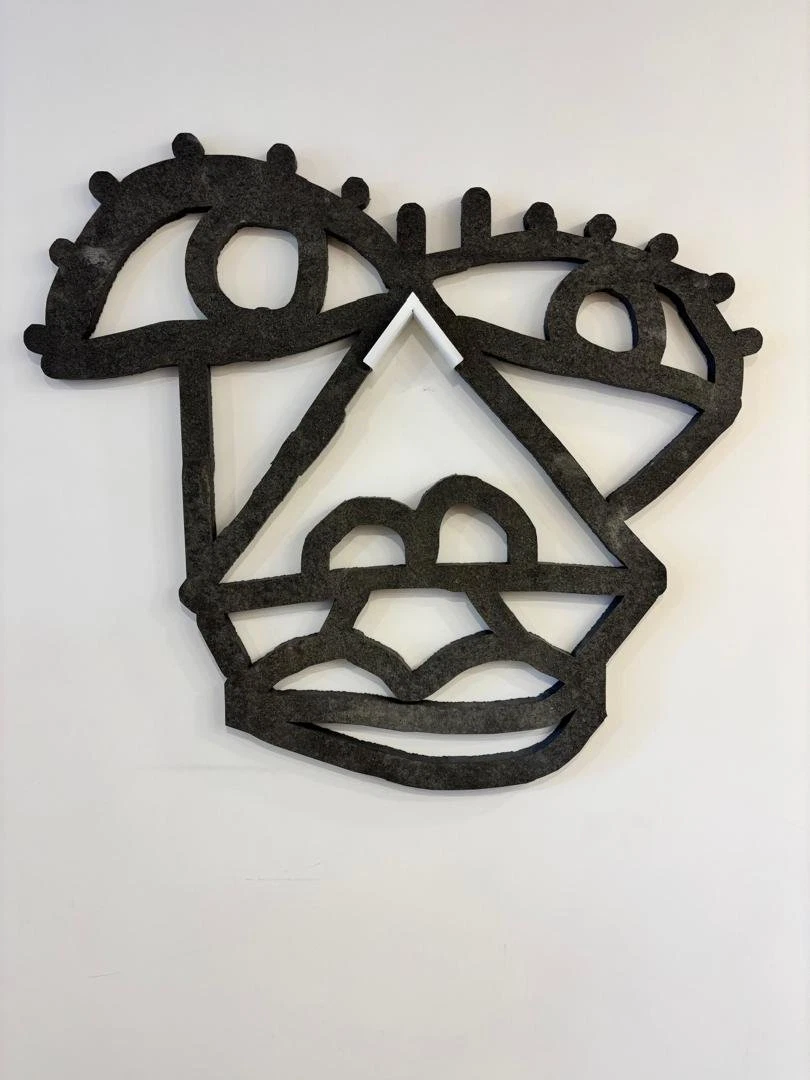 Rostro calado de piedra gris oscura, con ojos de pestañas, nariz triangular y labios, colgado de una pared blanca. Rostro en granito. Granito, 95 x 95 x 3 cm, 2025. Caracas. Nicolás Lapadula