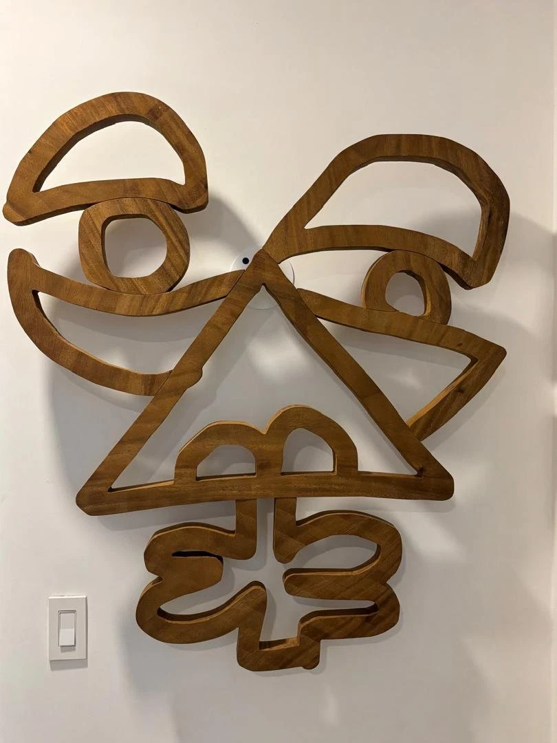 Rostro calado de madera con dos cejas curvas, nariz triangular y una forma de trébol bajo la boca, colgado de una pared blanca. Rostro en madera. Madera maciza, 100 x 95 x 4 cm, 2025. Caracas. Nicolás Lapadula
