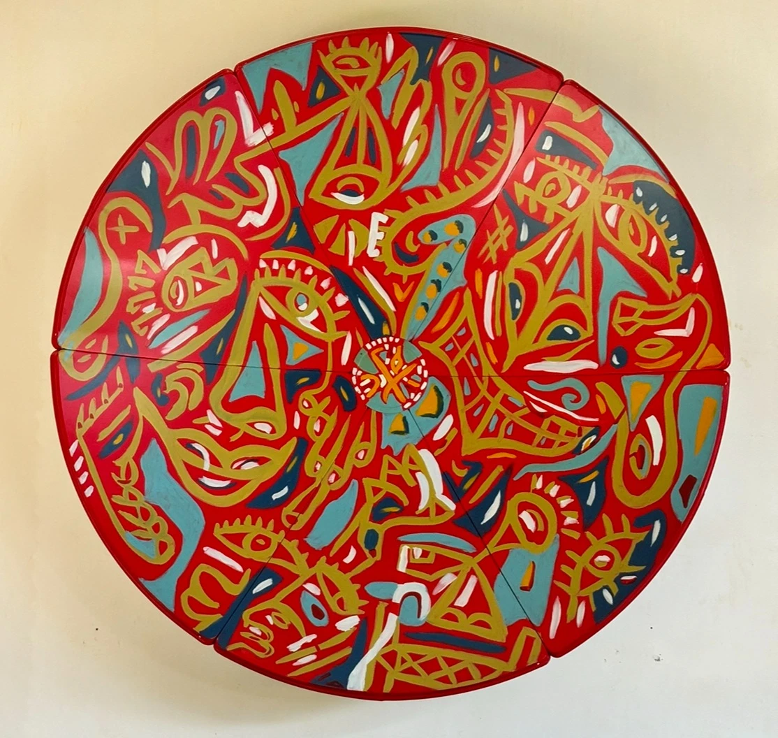 Disco circular de cuatro segmentos pintado en rojo, con trazos dorados y formas turquesa, azules y blancas que dibujan rostros. Sin título. Esmalte sobre aluminio, 240 cm de diámetro, 2024. Caracas. Nicolás Lapadula