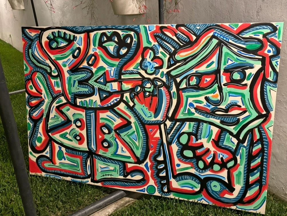 Dos figuras de rostro marcado en verde, rojo y azul con contornos negros gruesos sobre fondo crema, en un lienzo apoyado en un jardín. Sin título. Acrílico sobre lienzo, 180 x 120 cm, 2026. Caracas. Nicolás Lapadula