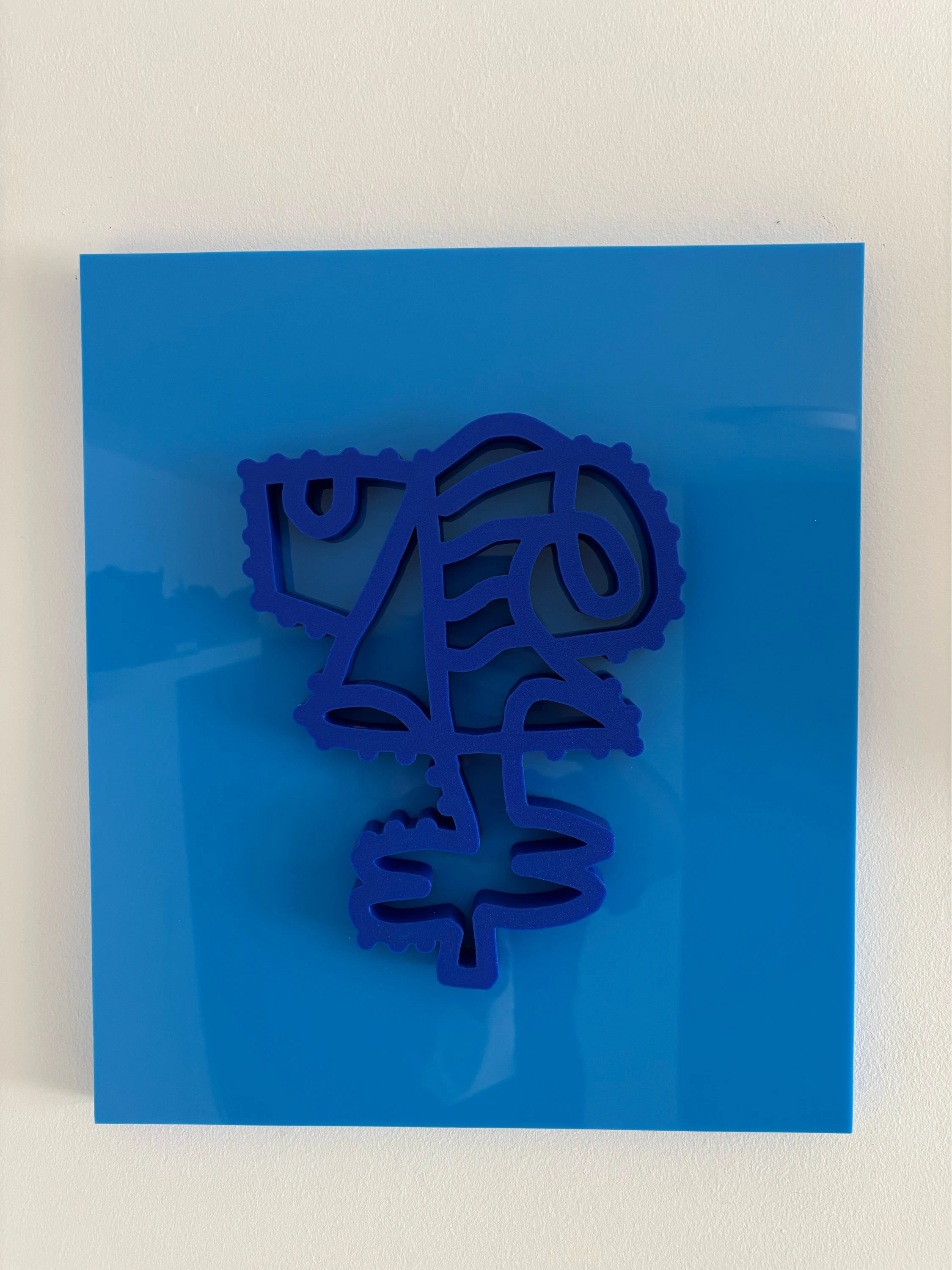 Rostro de trazo continuo en relieve azul oscuro, con borde de puntos y una forma de hojas abajo, sobre una placa de acrílico azul claro en una pared blanca. Sin título. Impresión 3D sobre acrílico, 35 x 40 cm, 2026. Caracas. Nicolás Lapadula
