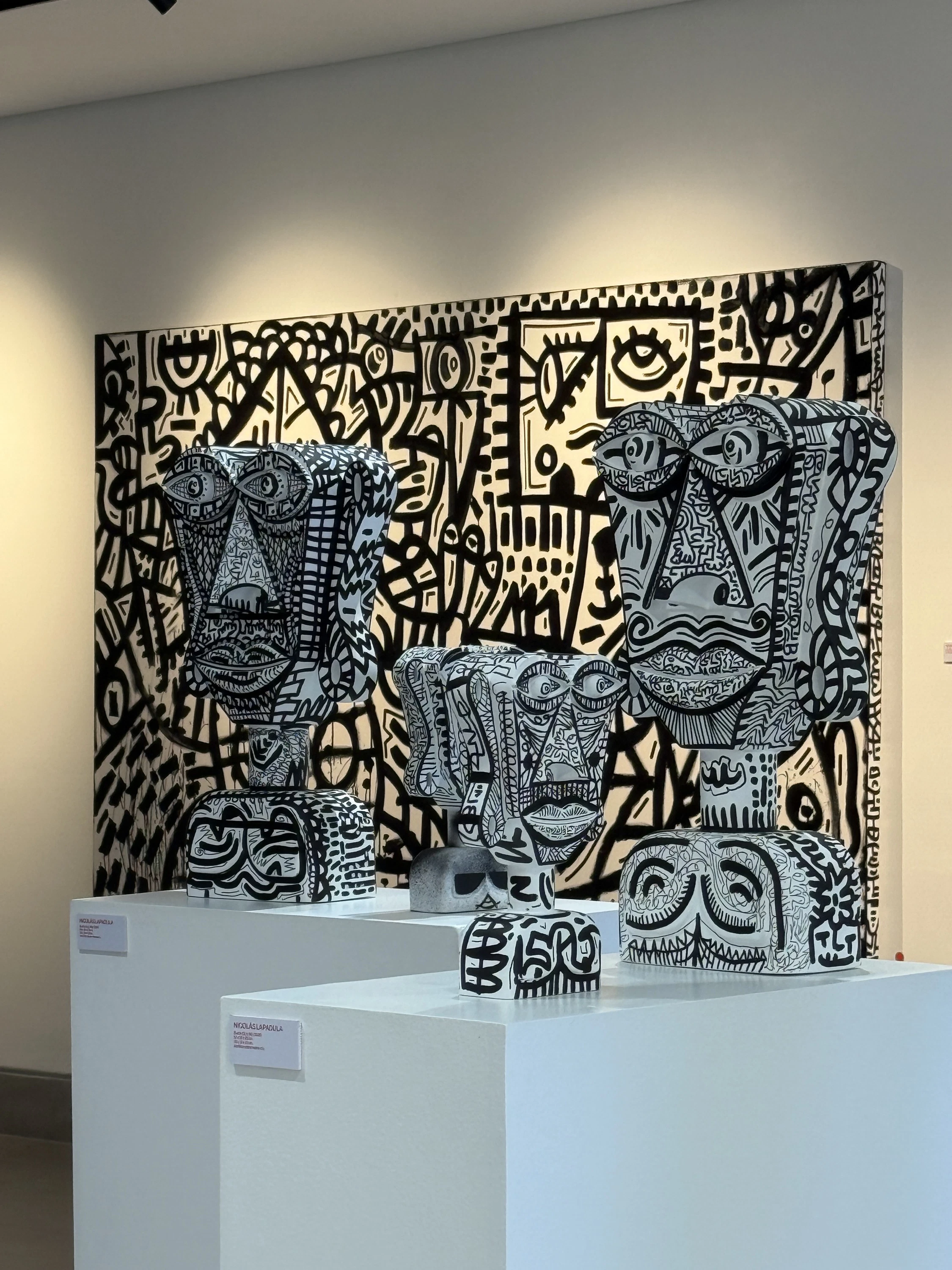 Sala de exposición con un gran lienzo de trazos negros sobre blanco y, delante, tres bustos blancos con dibujos negros sobre pedestales blancos. Sin título. Nicolás Lapadula