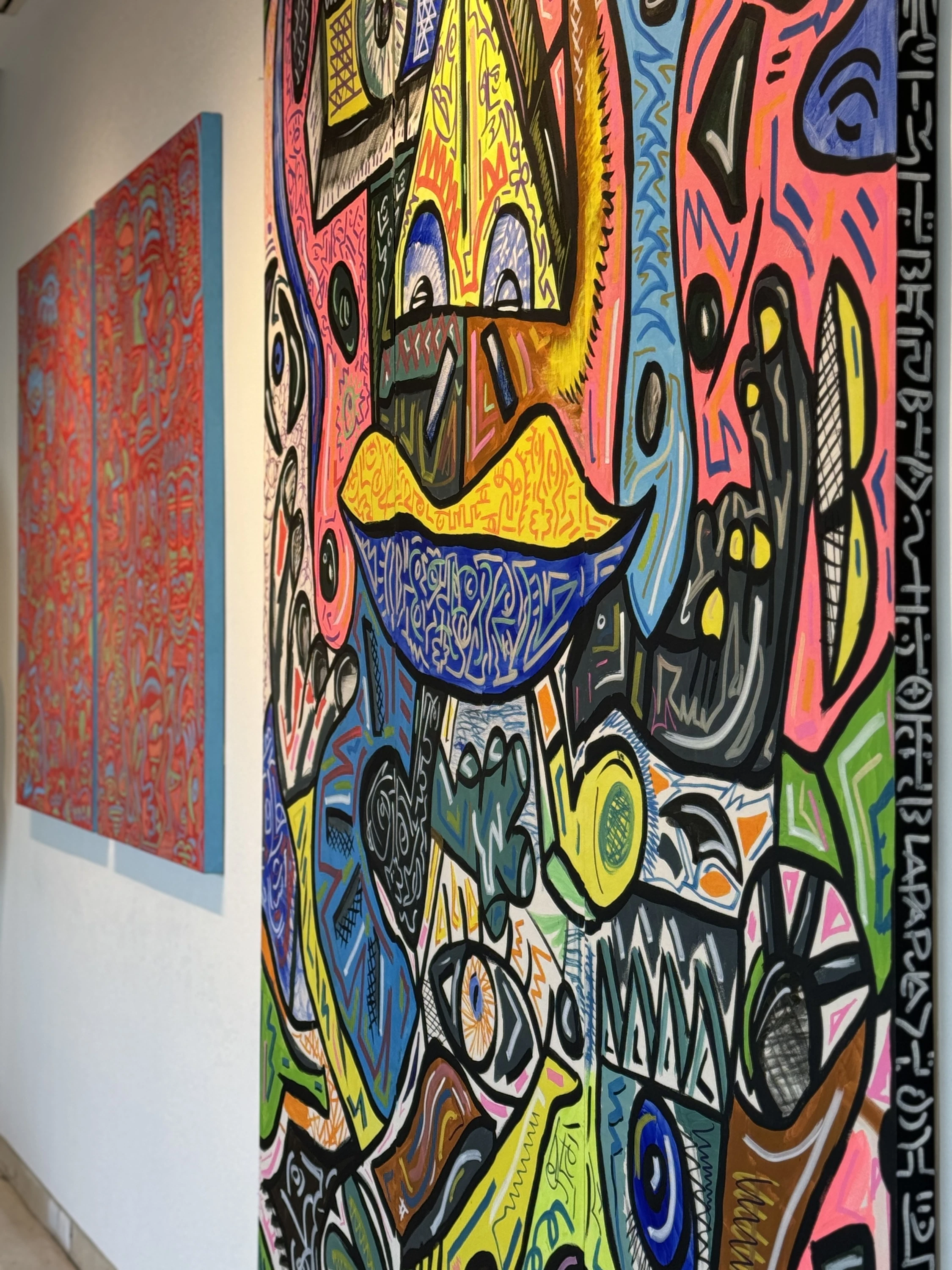 Vista de sala con un lienzo vertical de rosa, azul, verde y negro con una boca amarilla, y dos lienzos rojos al fondo. Sin título. Nicolás Lapadula