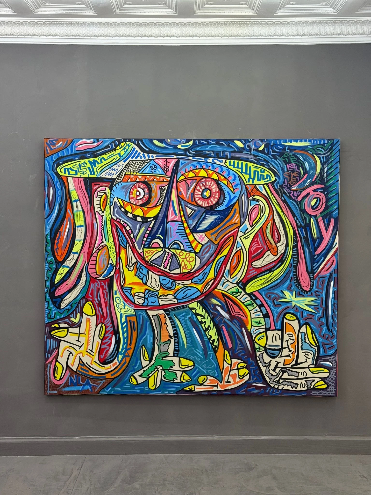 Lienzo de rostro azul y amarillo con ojos rosados y sonrisa roja, colgado en una pared gris dentro de una sala. Sin título. Nicolás Lapadula