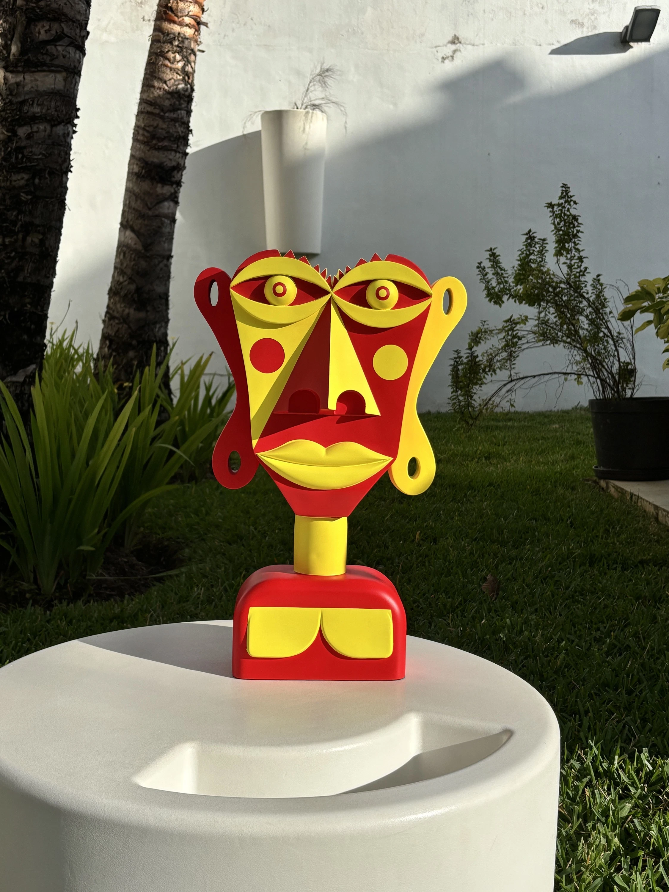 Busto escultórico de rostro rojo y amarillo con ojos saltones, lunares y orejas laterales, sobre una base redonda blanca en un jardín. Sin título. Nicolás Lapadula