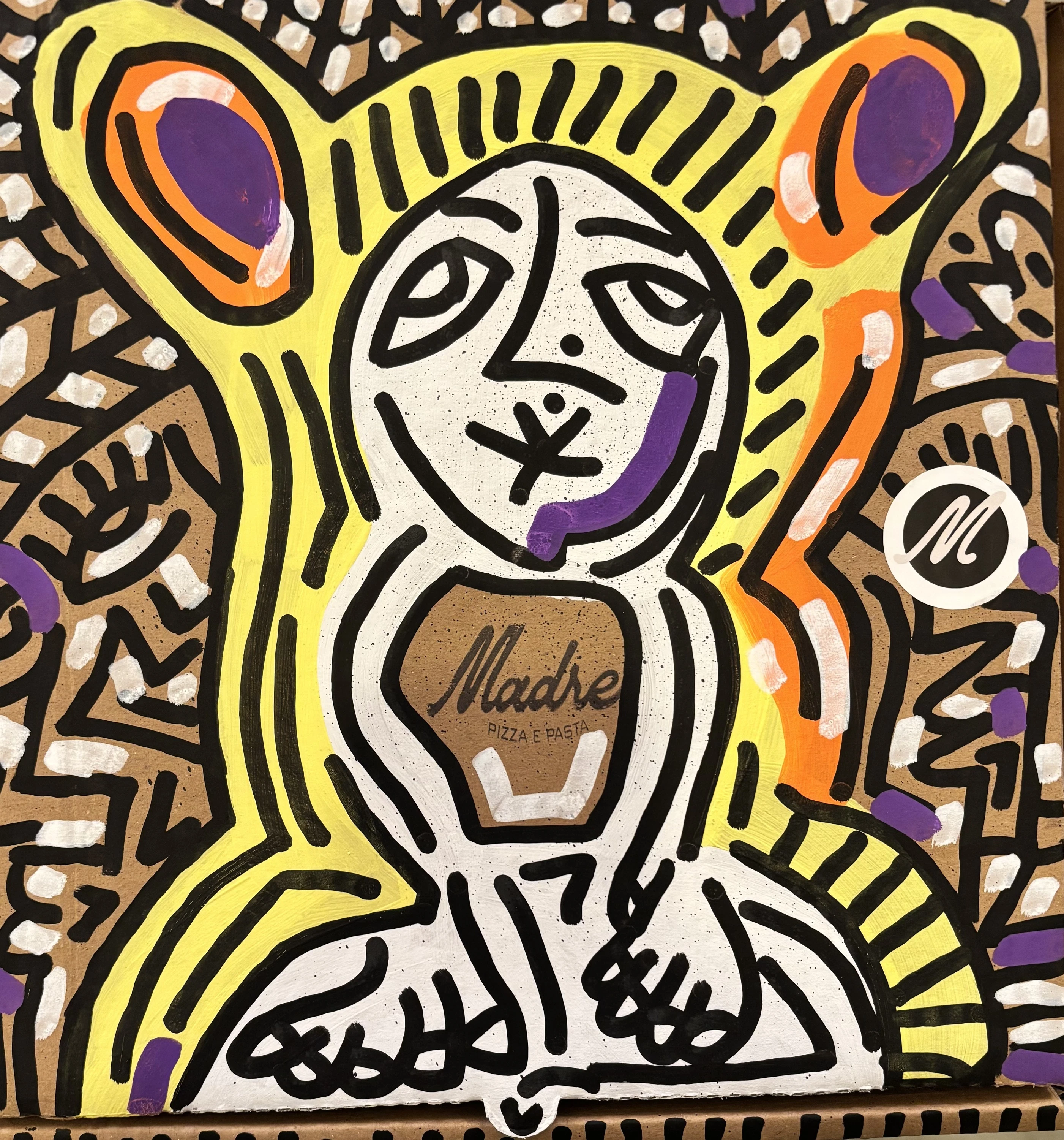Figura de rostro blanco con halo amarillo y dos óvalos naranja y morado a los lados, en trazos negros sobre una caja de cartón. Sin título. Acrílico sobre cartón, 40 x 40 cm, 2026. Caracas. Nicolás Lapadula
