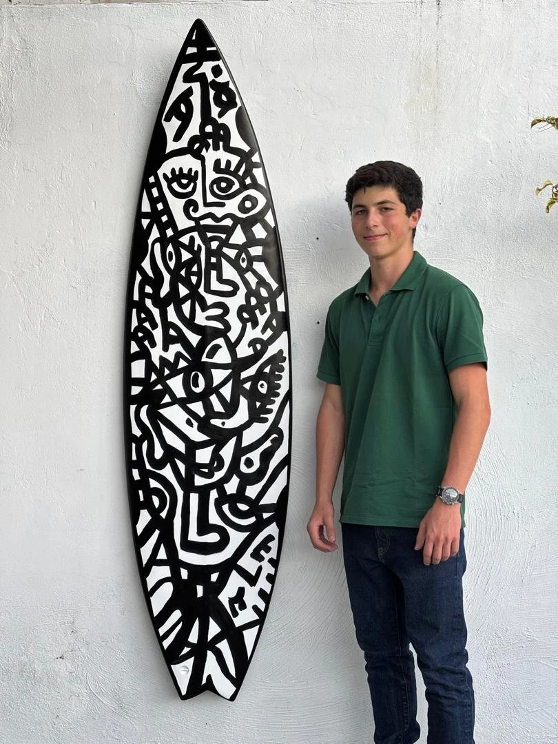 Tabla de surf colgada de una pared blanca, decorada con rostros y líneas negras sobre blanco; a su lado hay un joven con polo verde. Surfboard. Acrílico y poliuretano sobre fibra de vidrio, 50 x 183 cm, 2025. Caracas. Nicolás Lapadula